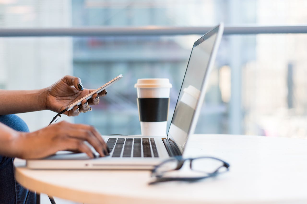 Close-up of hands using a laptop and phone with coffee on a modern office desk.